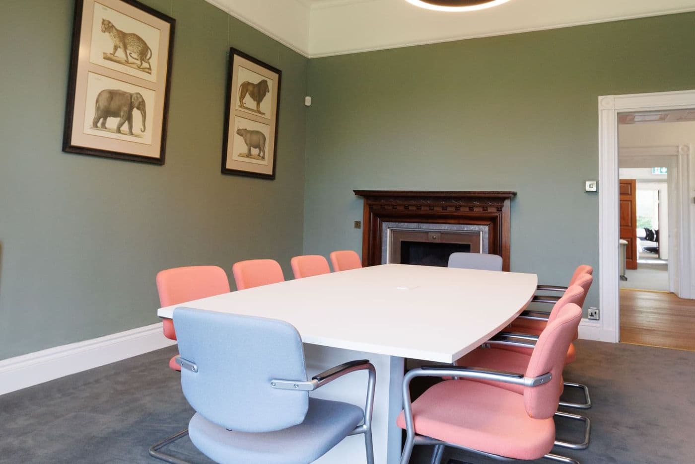 Bright conference room with white table, pink and blue chairs, green walls, framed animal prints and a decorative wooden fireplace.