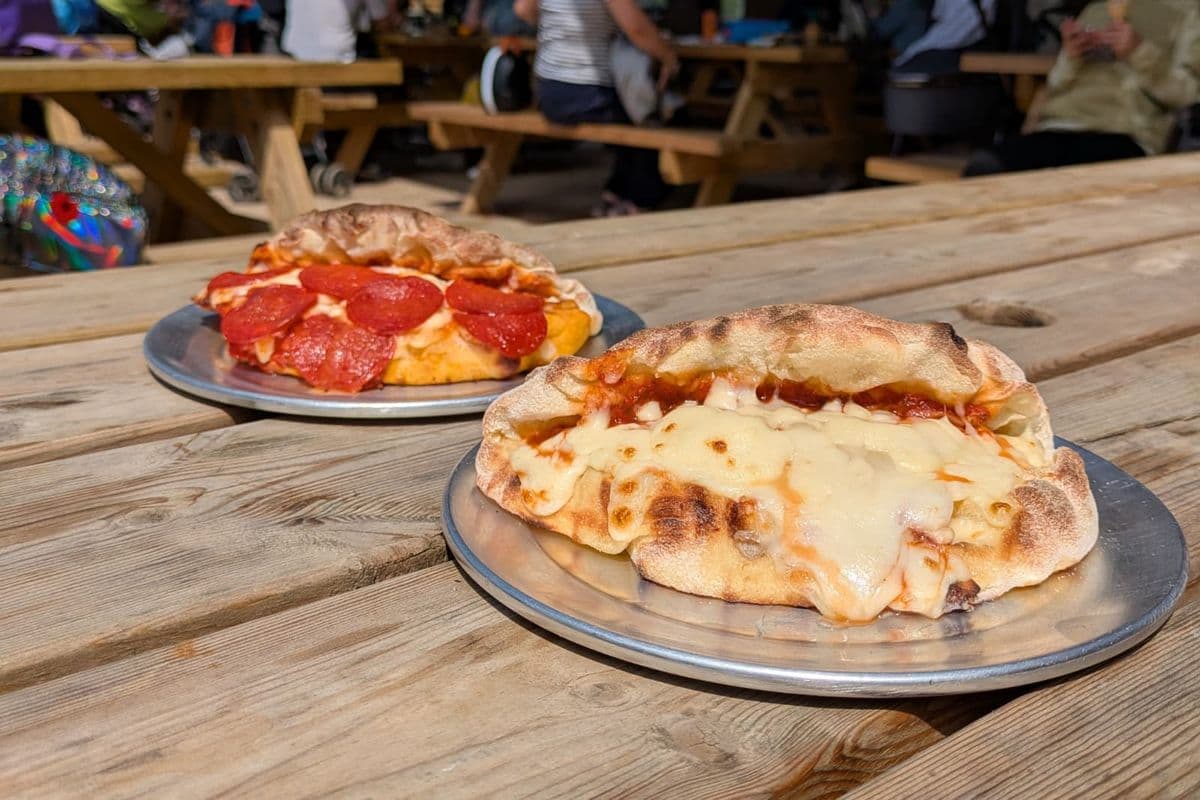 Two folded pizzas on metal plates sit on a wooden picnic table. One has pepperoni, and the other is topped with melted cheese.